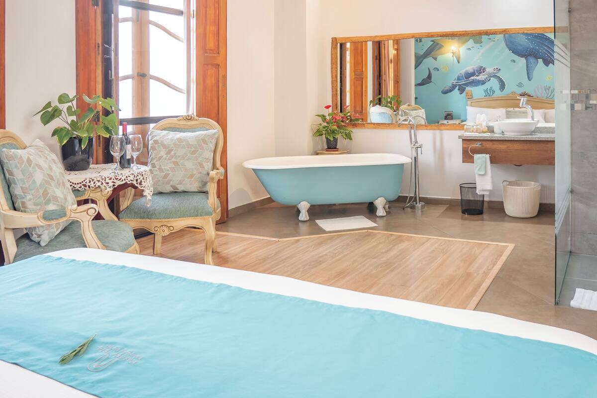 A bedroom with a bed, a chair, a bathtub, and a bathroom with a mirror and tiles.