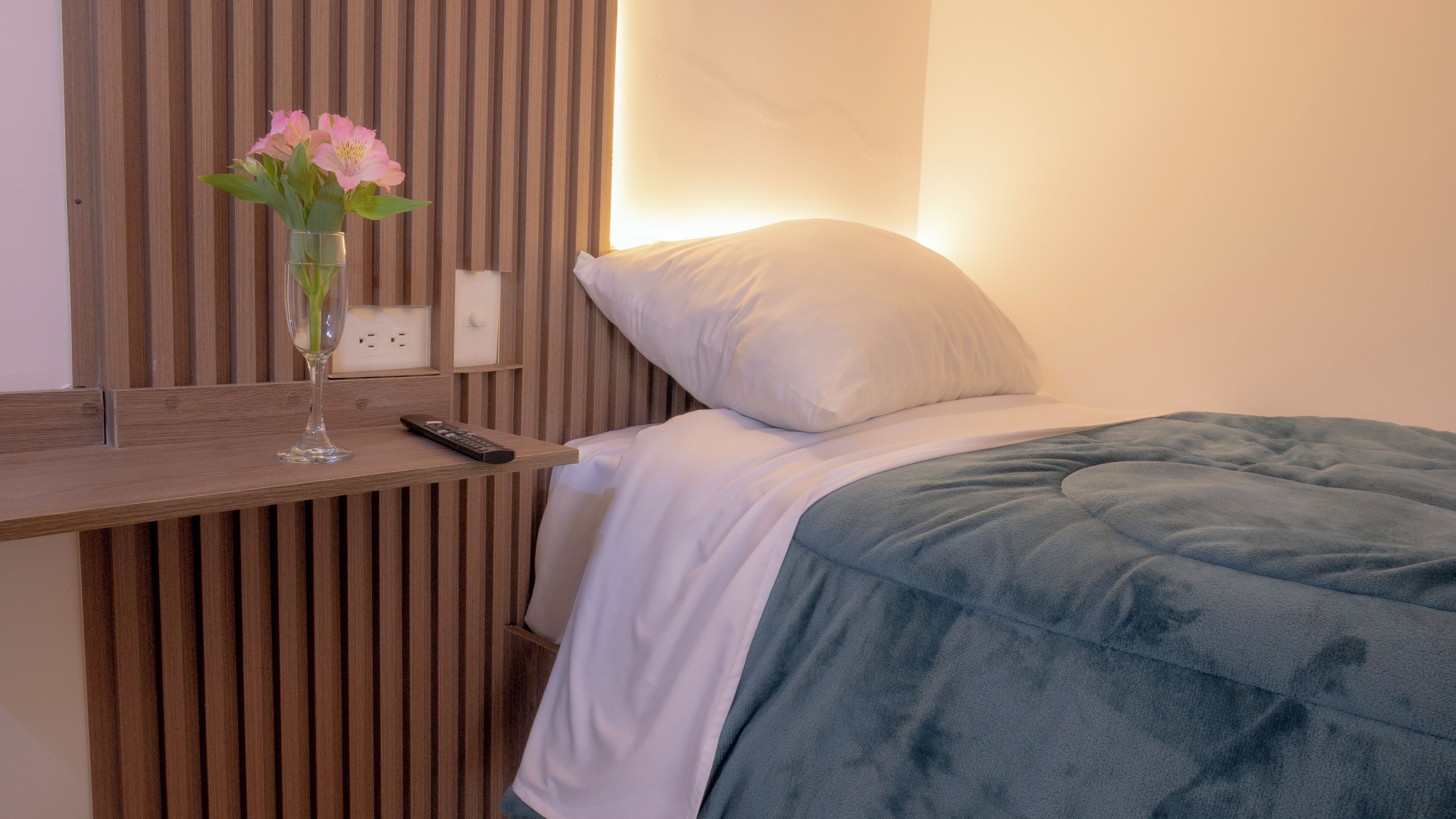 A hotel room with a wooden slat wall, a bedside table with a vase of flowers, a remote control, and a bed with a white pillow and blue blanket.