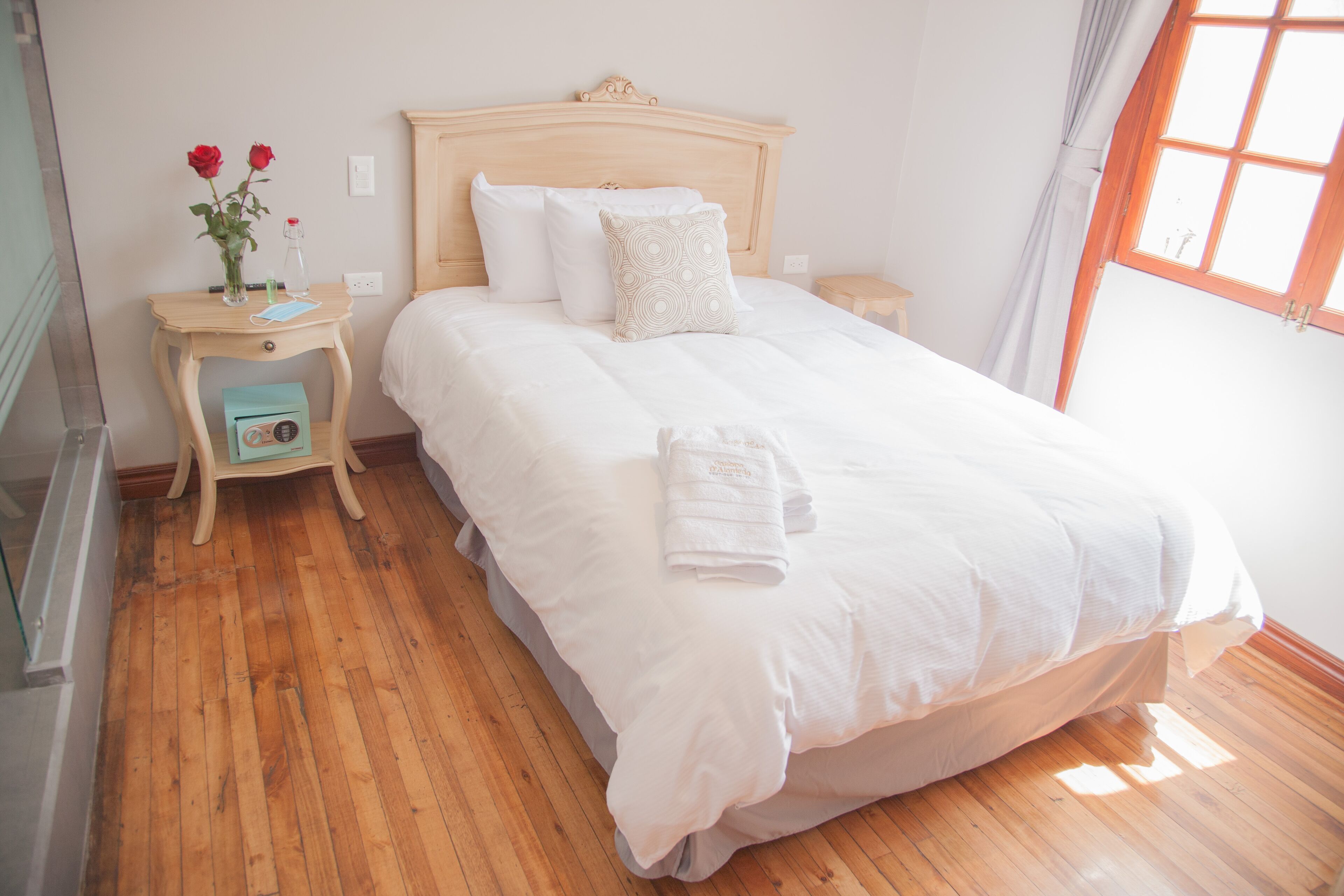 A neatly made bed with white linens, a wooden headboard, and a bedside table with a vase of red roses.