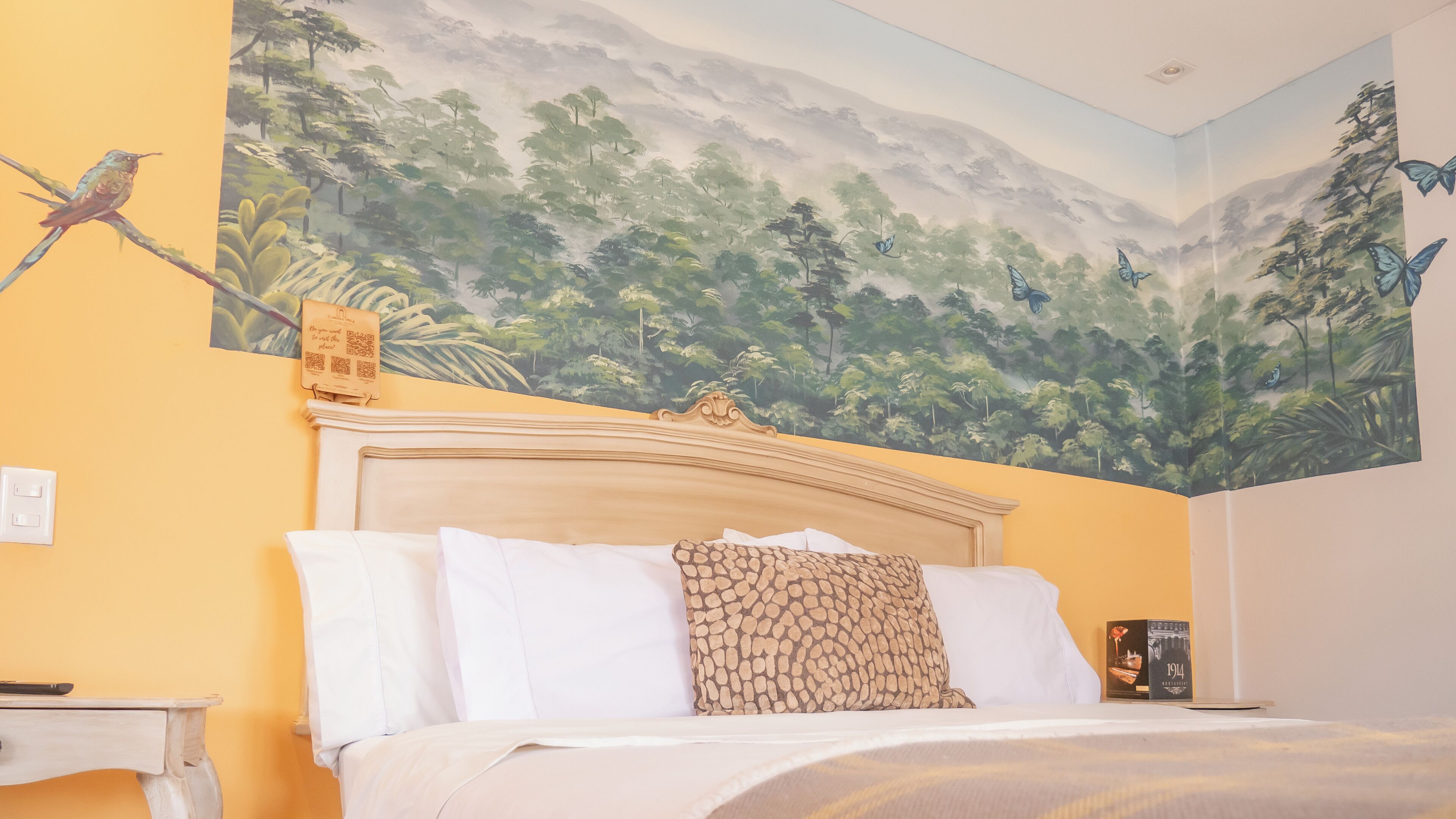 Un dormitorio con un mural de un paisaje forestal, una cama con sábanas blancas y un cojín estampado, y una mesita de noche con un libro y un cartel con un código QR.