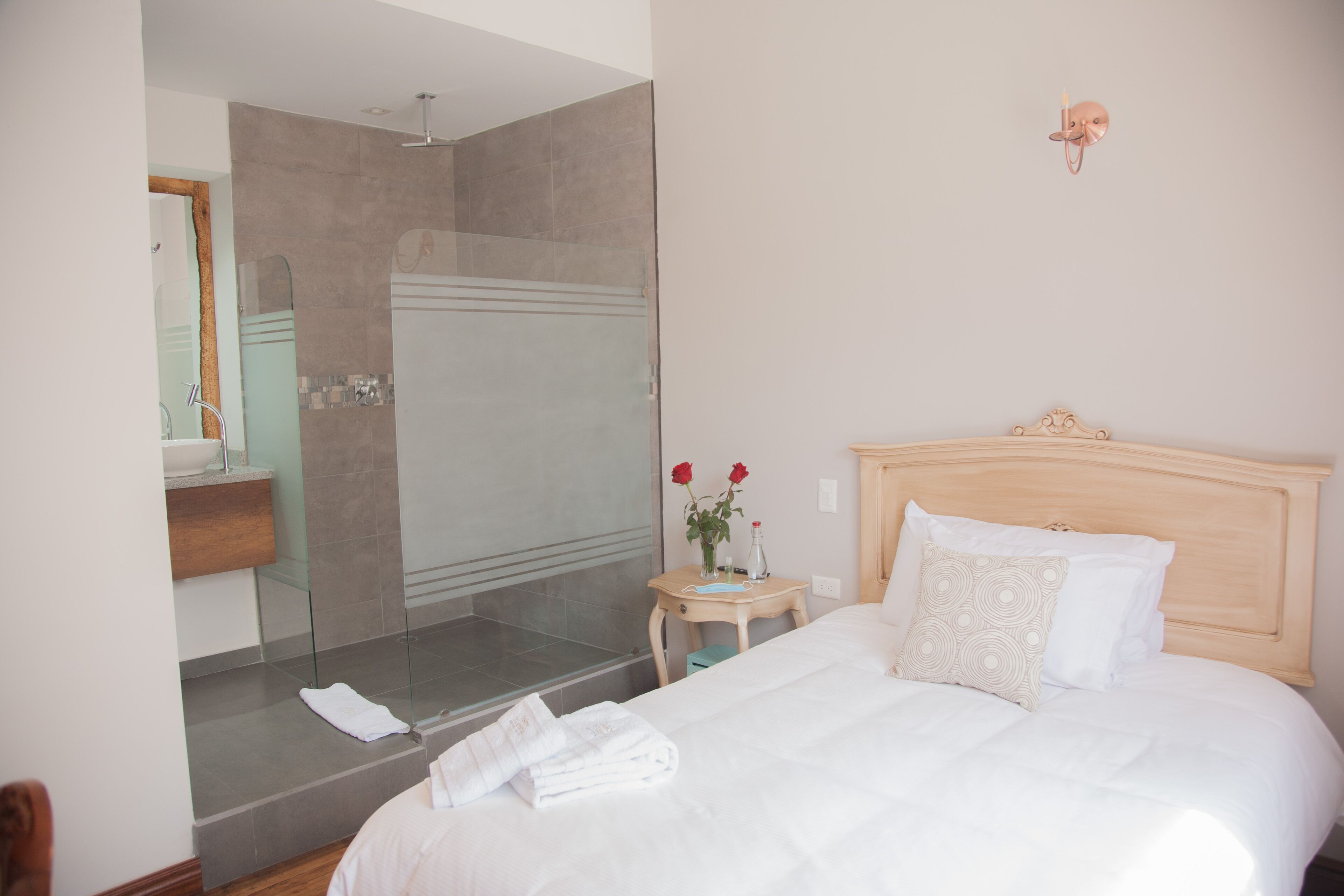 A hotel room with a large bed, a bedside table with a vase of flowers, a mirror, and a shower area.