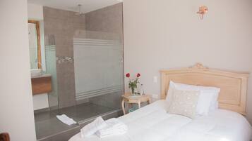 A hotel room with a large bed, a bedside table with a vase of flowers, a mirror, and a shower area.