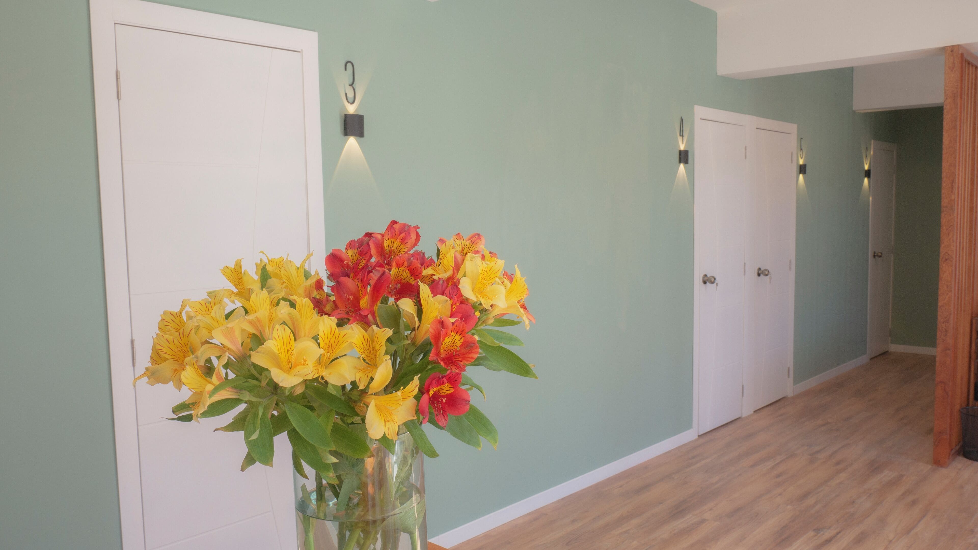 Un couloir avec un vase de fleurs, des portes blanches et des lampes fixées au mur.