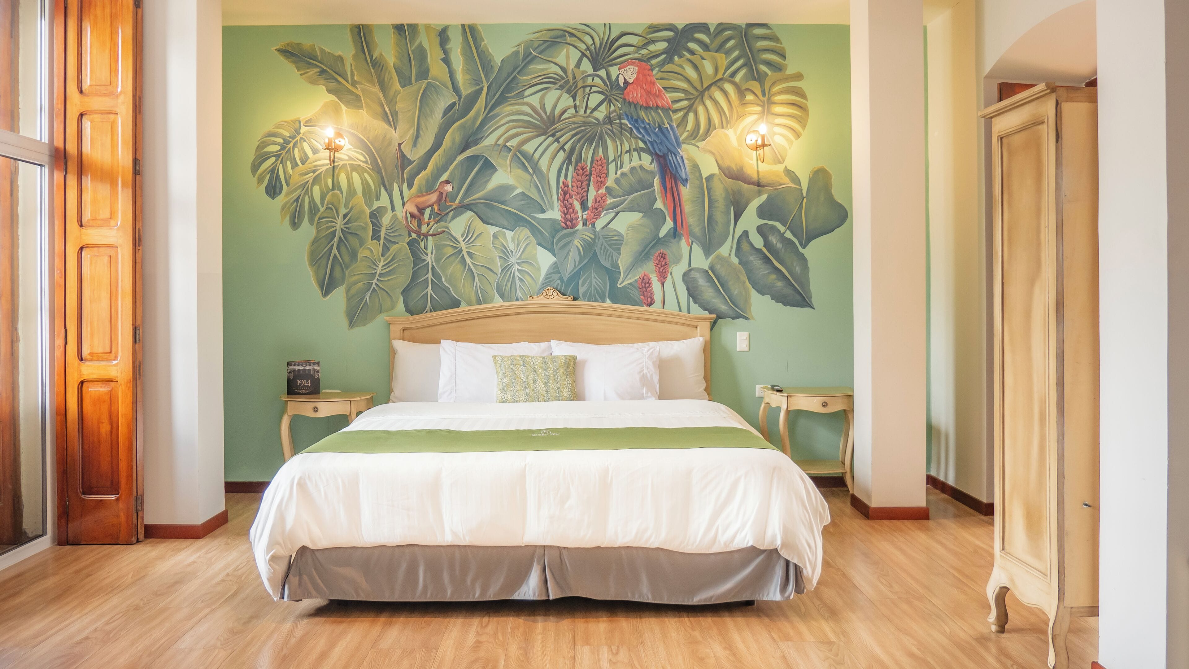 A bedroom with a large bed, a nightstand, a wooden wardrobe, and a tropical mural on the wall.