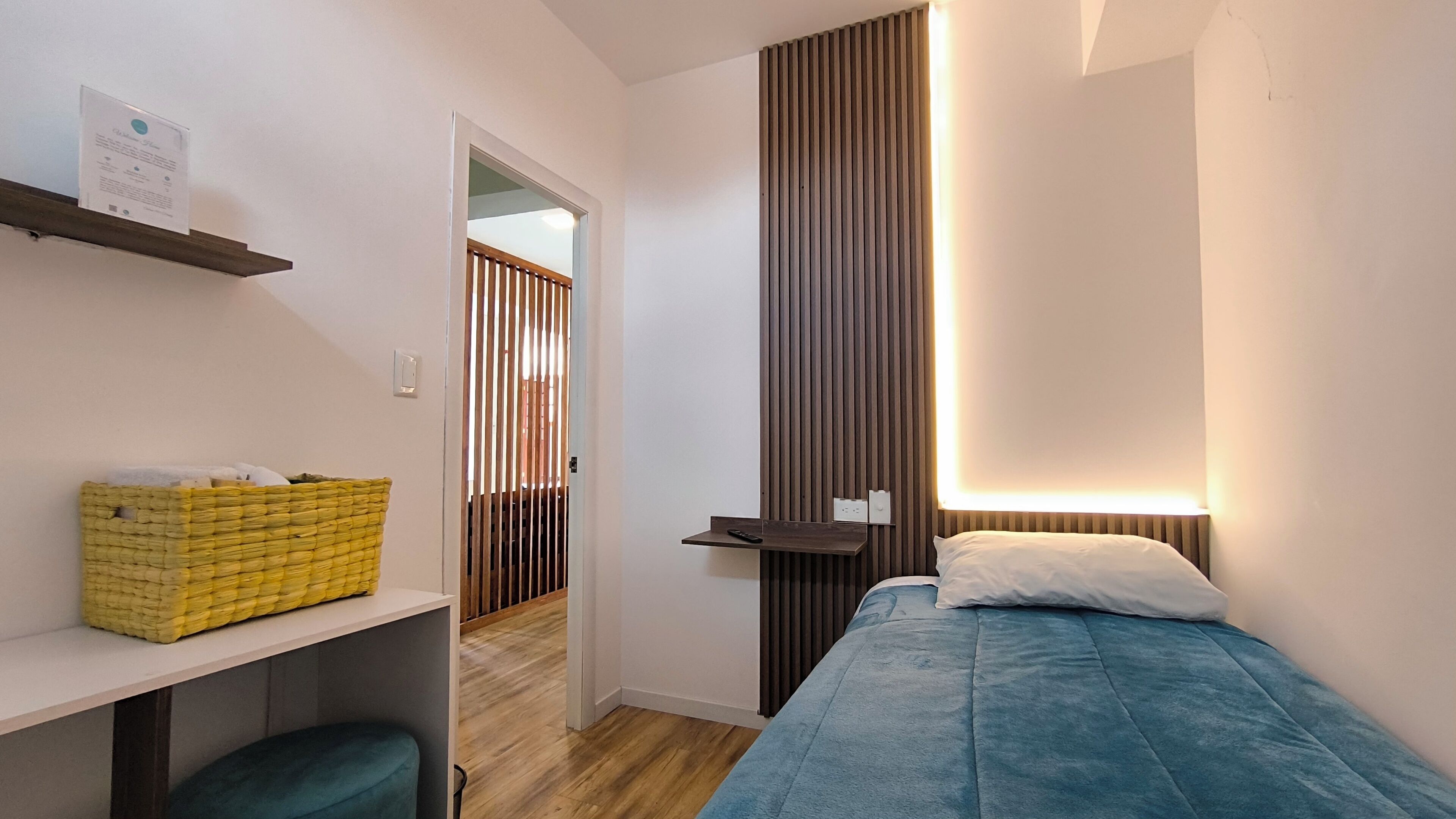 Een slaapkamer met een bed, een nachtkastje, een wandlamp, een houten vloer en een deur naar een andere kamer.