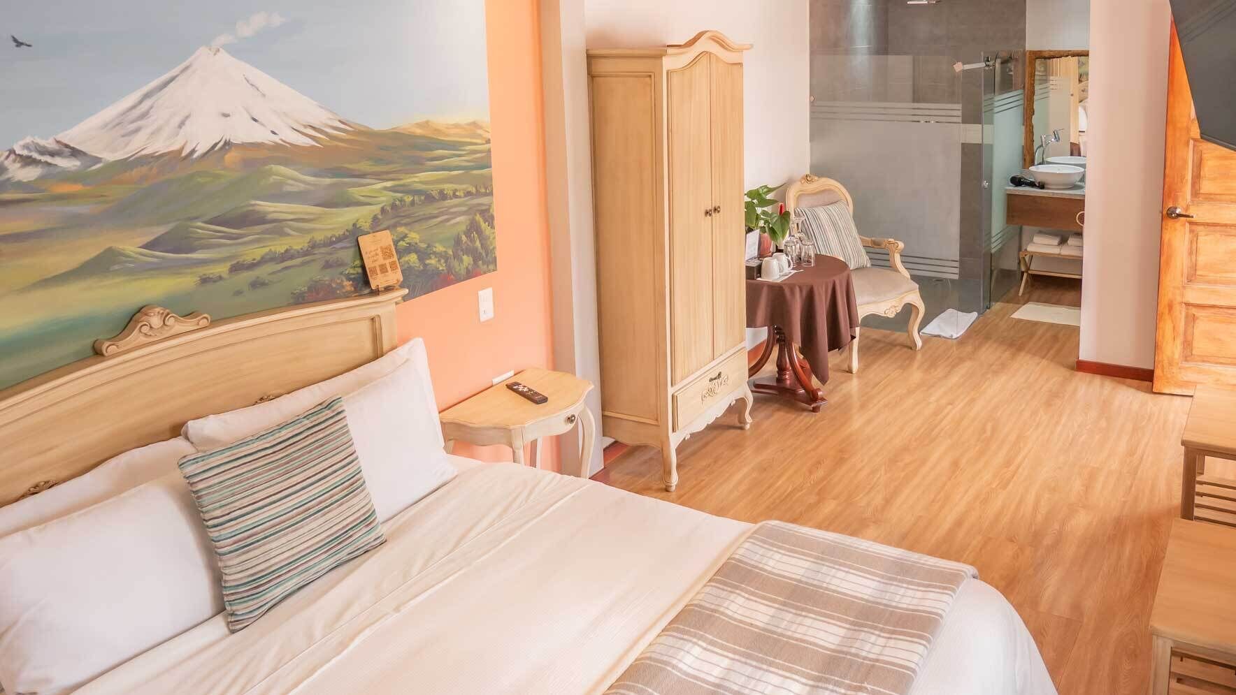 A hotel room with a bed, a wooden wardrobe, a dining table with chairs, a bathroom with a mirror, and a painting of a mountain.