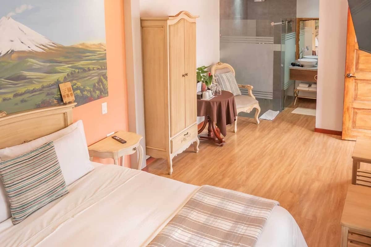 A hotel room with a bed, a wooden wardrobe, a dining table with chairs, a bathroom with a mirror, and a painting of a mountain.