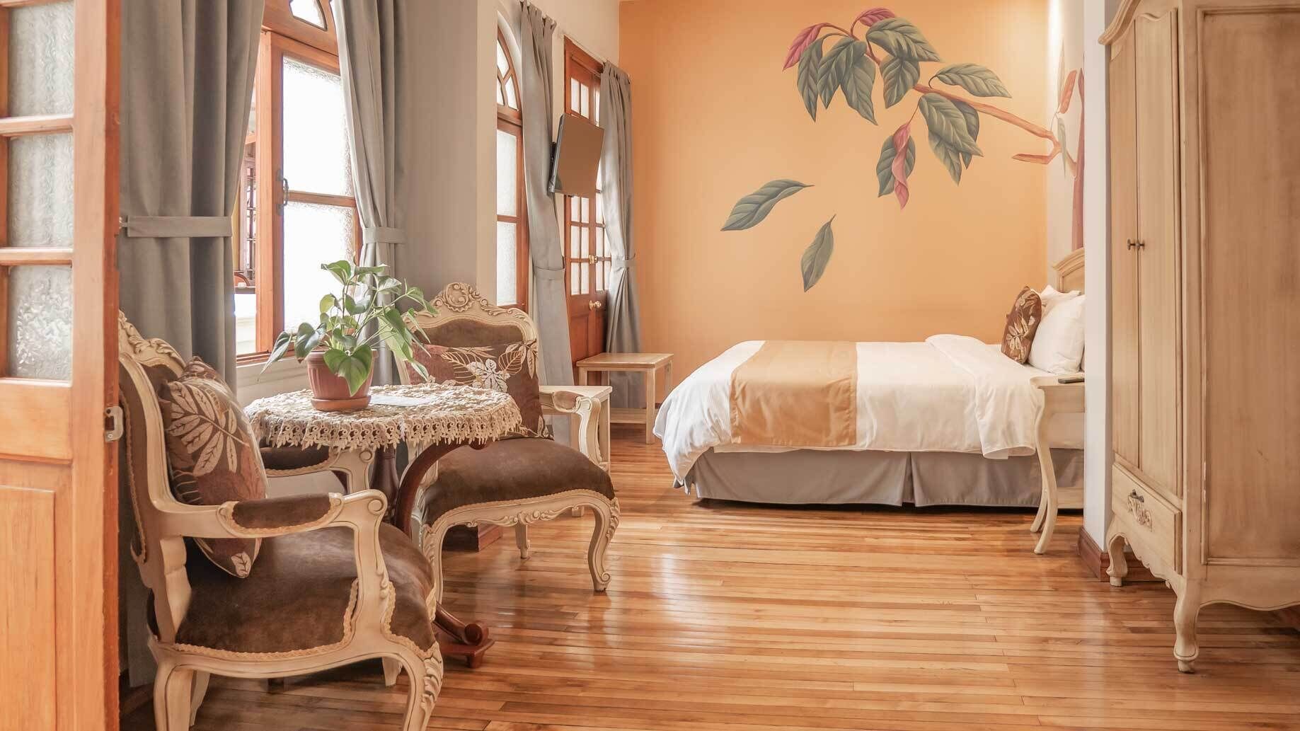 A room with a bed, a table with chairs, a potted plant, and a wall with a leaf mural.