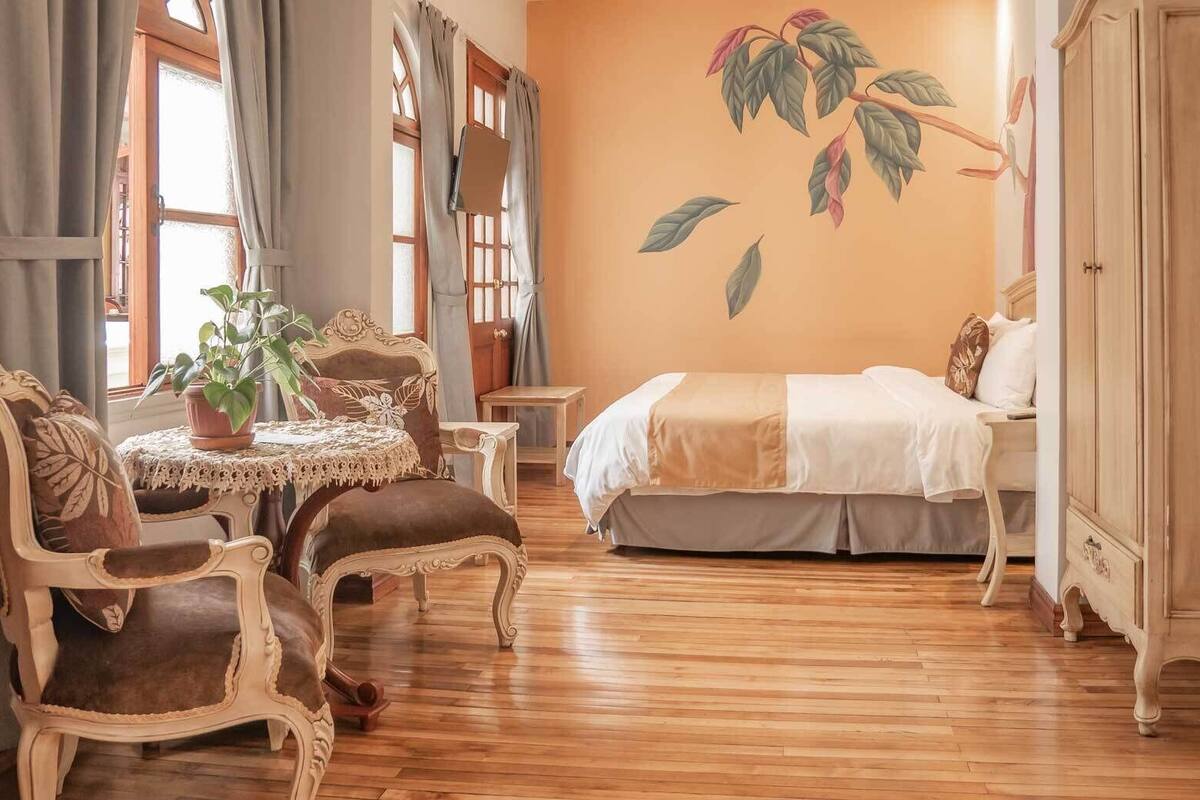 A room with a bed, a table with chairs, a potted plant, and a wall with a leaf mural.