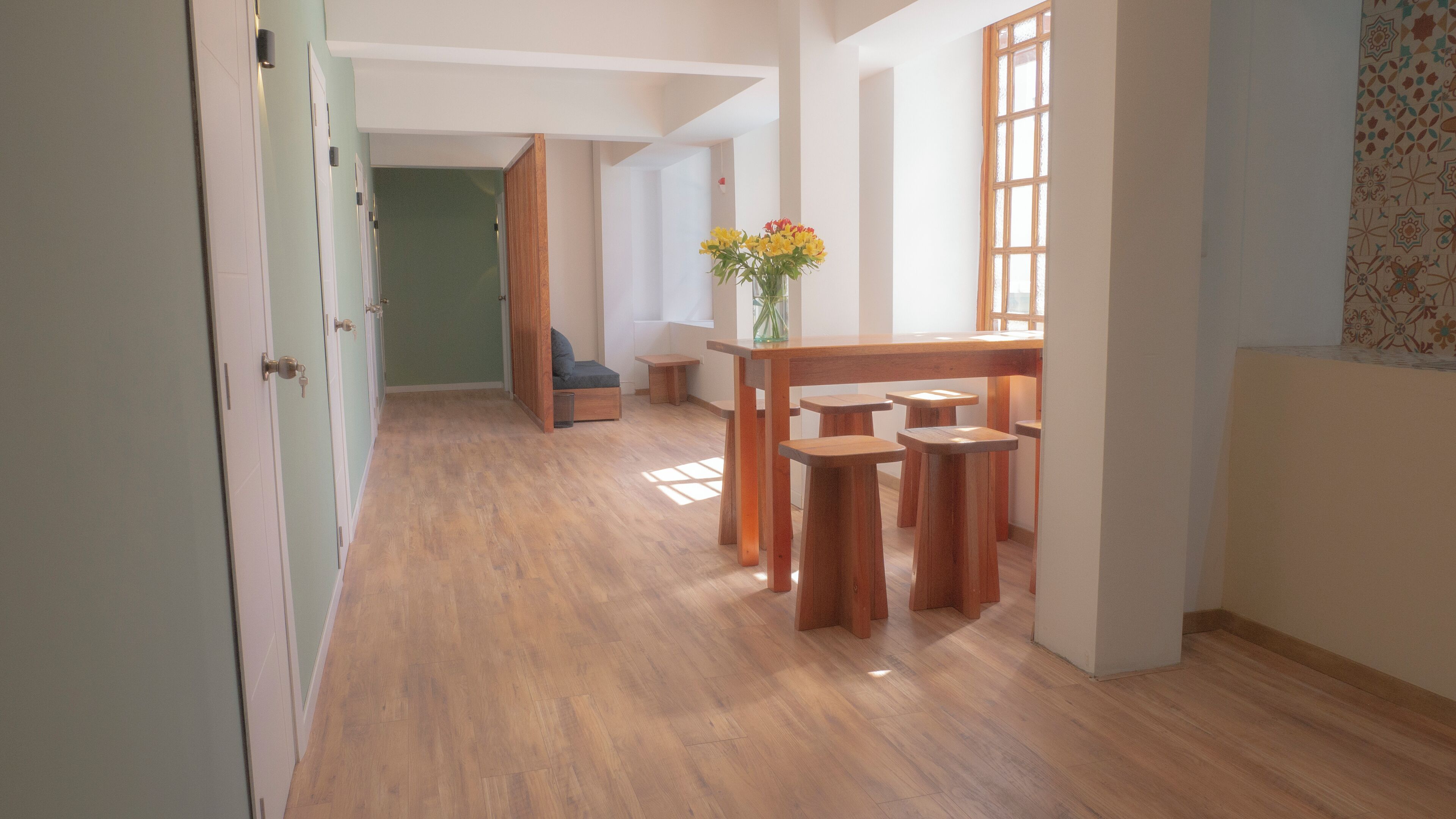 A hallway with wooden flooring, a wooden table with stools, a vase with flowers, and a blue chair.