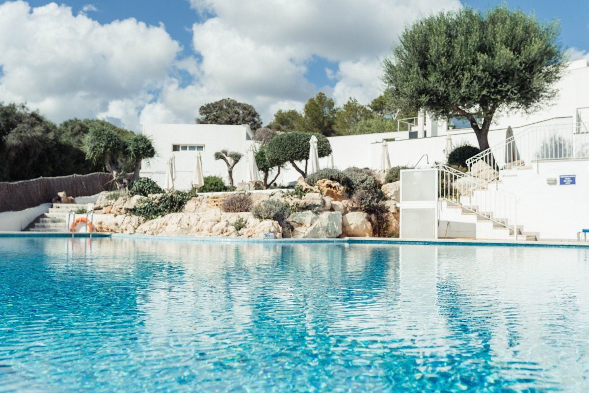 A swimming pool with clear blue water, surrounded by white buildings, trees, and a rocky landscape.