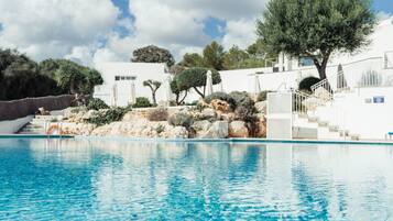 A swimming pool with clear blue water, surrounded by white buildings, trees, and a rocky landscape.