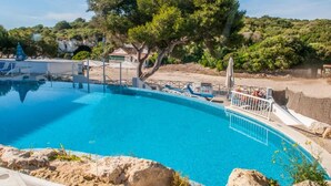 Una piscina azul cristalina con tobogán, rodeada de vegetación y árboles.