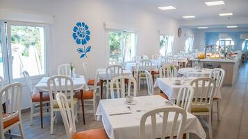 A dining area with white tables and chairs, a blue floral wall decoration, and a breakfast bar with fruit and beverages.