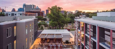 A rooftop pool area with lounge seating and a covered patio, surrounded by modern buildings and a sunset sky.