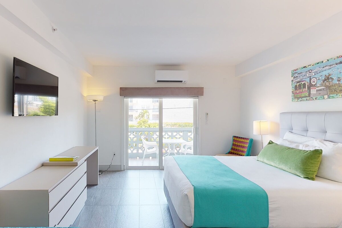 A bedroom with a bed, a desk, a chair, a TV, and a balcony with a view.