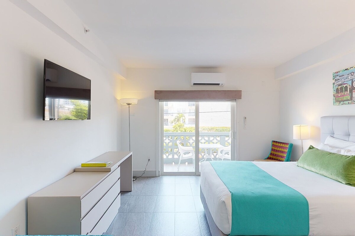 A bedroom with a bed, a desk, a chair, a TV, and a balcony with a view.
