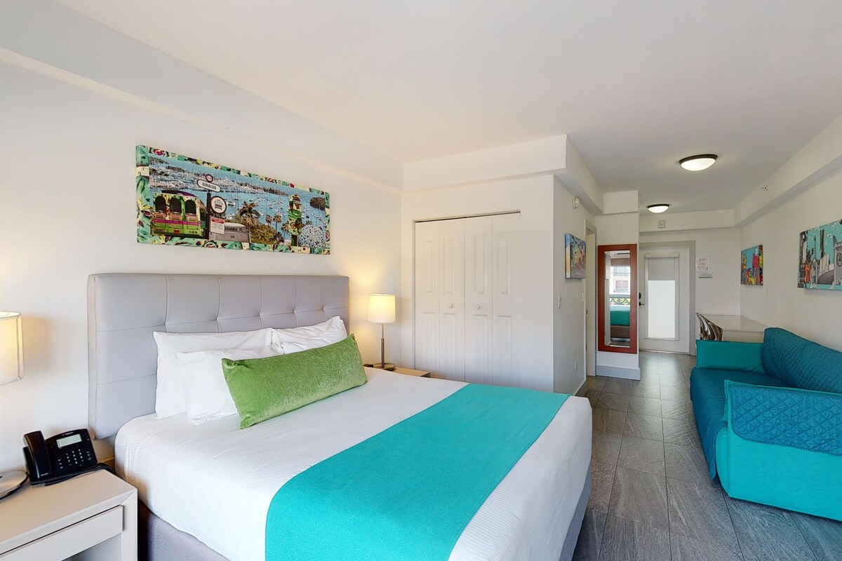 A hotel room with a bed, a desk, a phone, a door, and a hallway leading to another room.