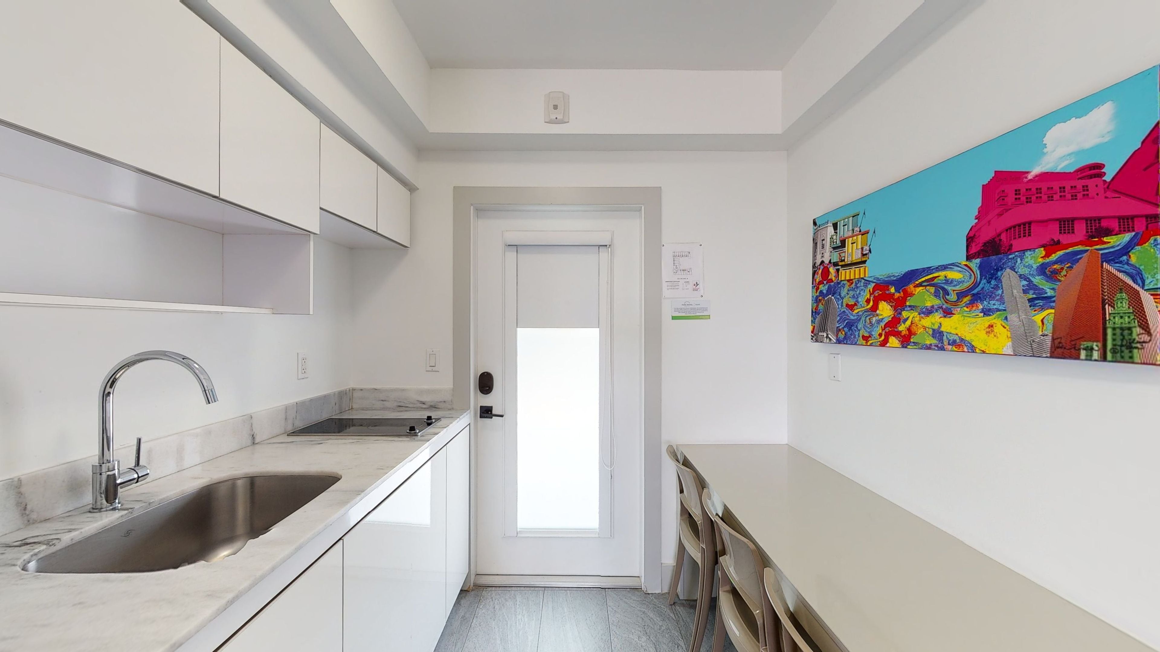 Una cocina estrecha con un lavamanos de acero inoxidable, armarios blancos y una puerta que da a otra habitación.