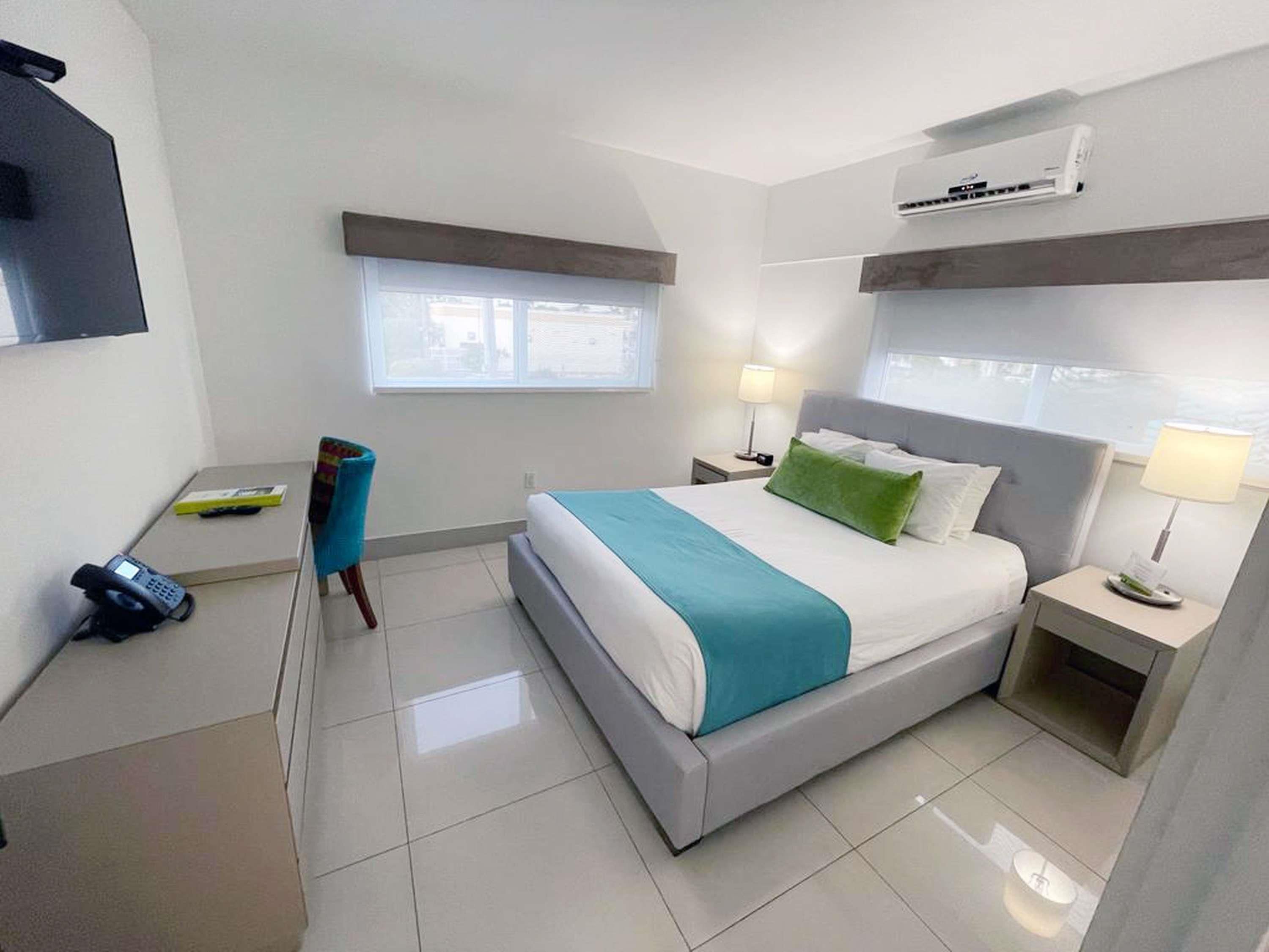 A hotel room with a bed, bedside tables, a chair, a desk, a phone, and an air conditioning unit.