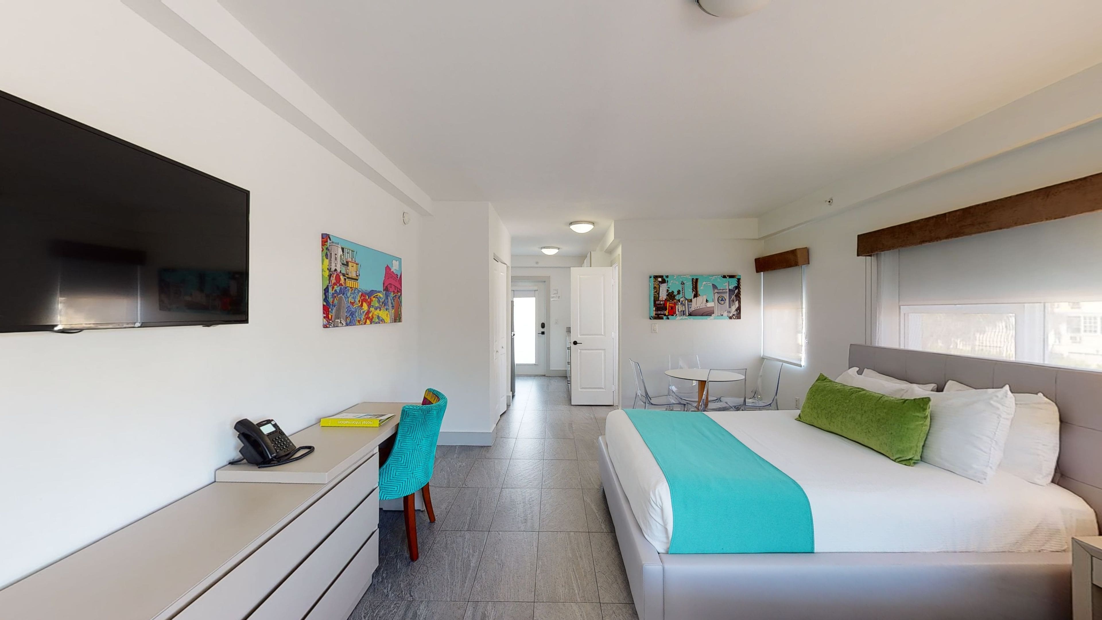 A hotel room with a bed, a desk, a chair, a TV, and artwork on the walls.