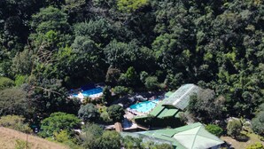 An aerial view of a resort with a swimming pool surrounded by dense forest, a covered area with a green roof, and a hillside. - Hostería Río Escondido (San Jerónimo)