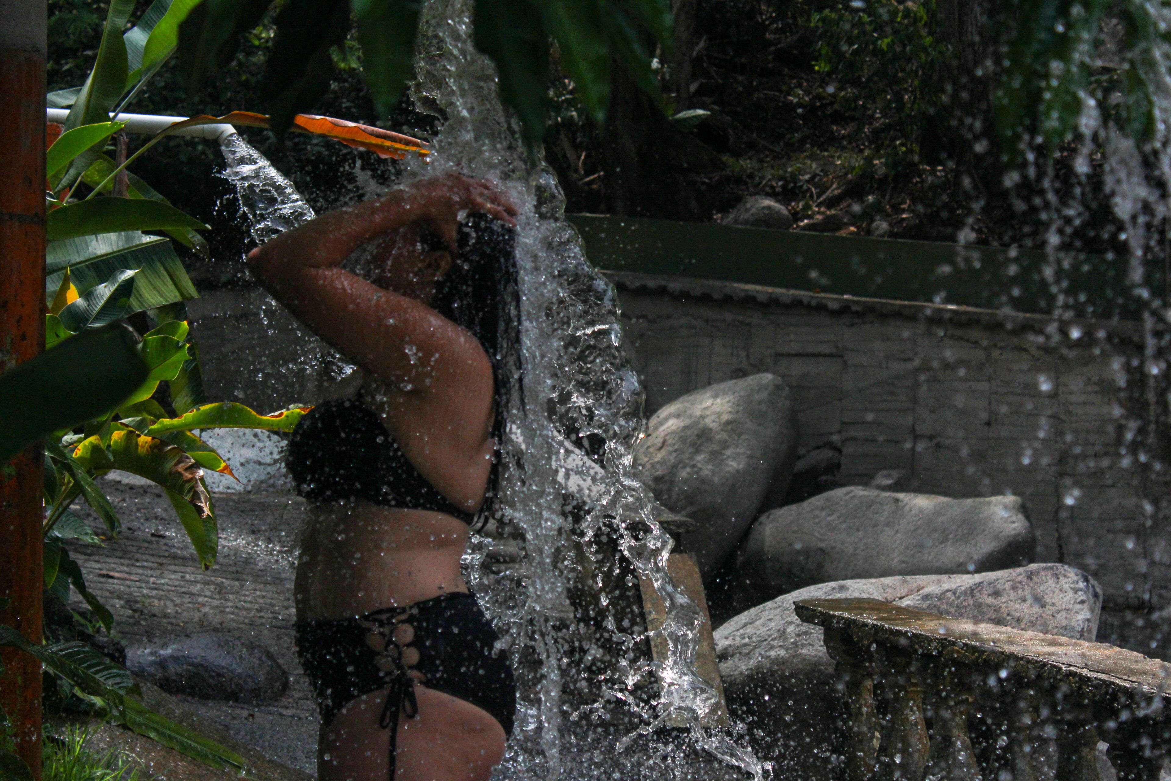 A person in a black swimsuit standing near a water feature with rocks and greenery in the background.