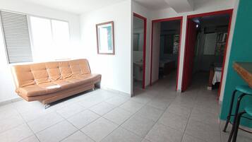 Un soggiorno con un divano marrone, un quadro incorniciato appeso al muro e un pavimento piastrellato. Si apre una porta che dà su un bagno e una zona cucina.