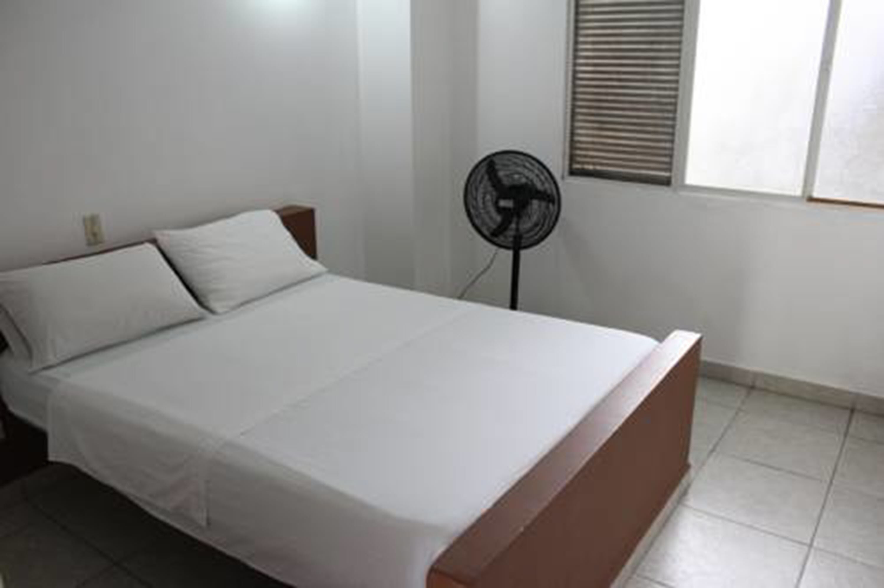 A single-bed room with a wooden headboard, a standing fan, and a window with blinds.