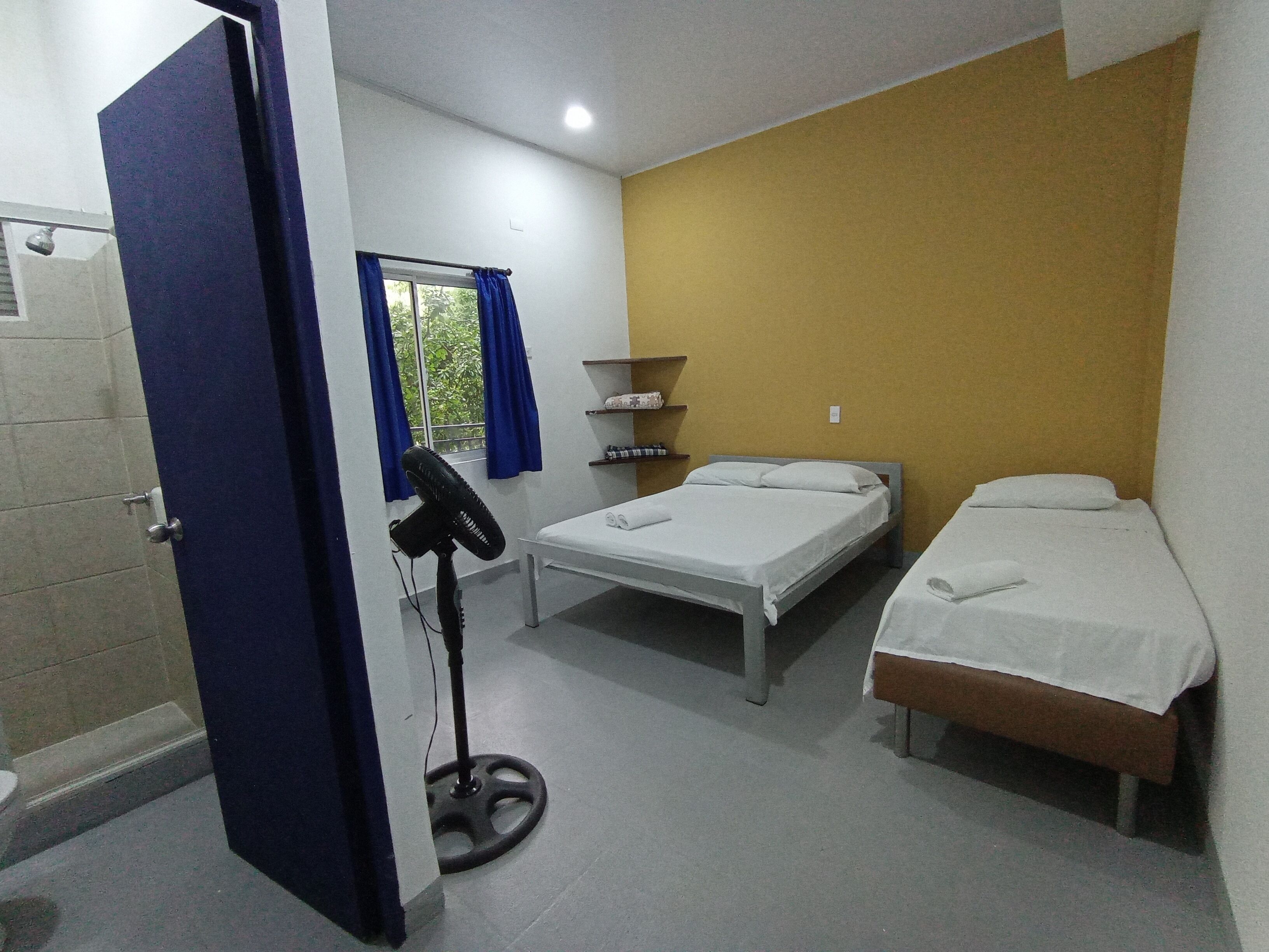 A room with two single beds, a standing fan, a shelf with toiletries, and a window with blue curtains.