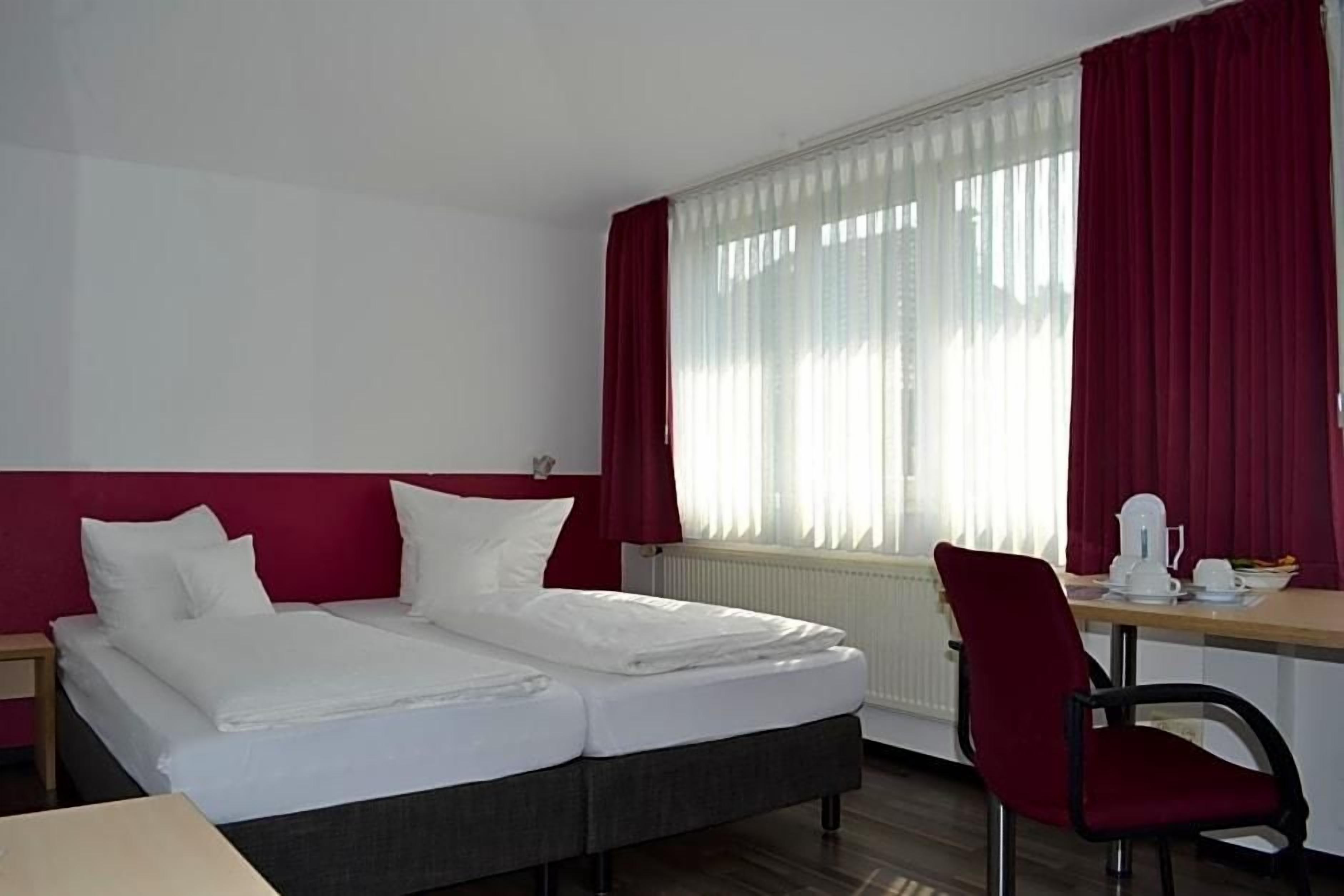 Una habitación de hotel con cama, escritorio, silla y ventana con cortinas.