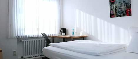 A hotel room with a bed, a desk with a chair, a radiator, and a window with curtains.