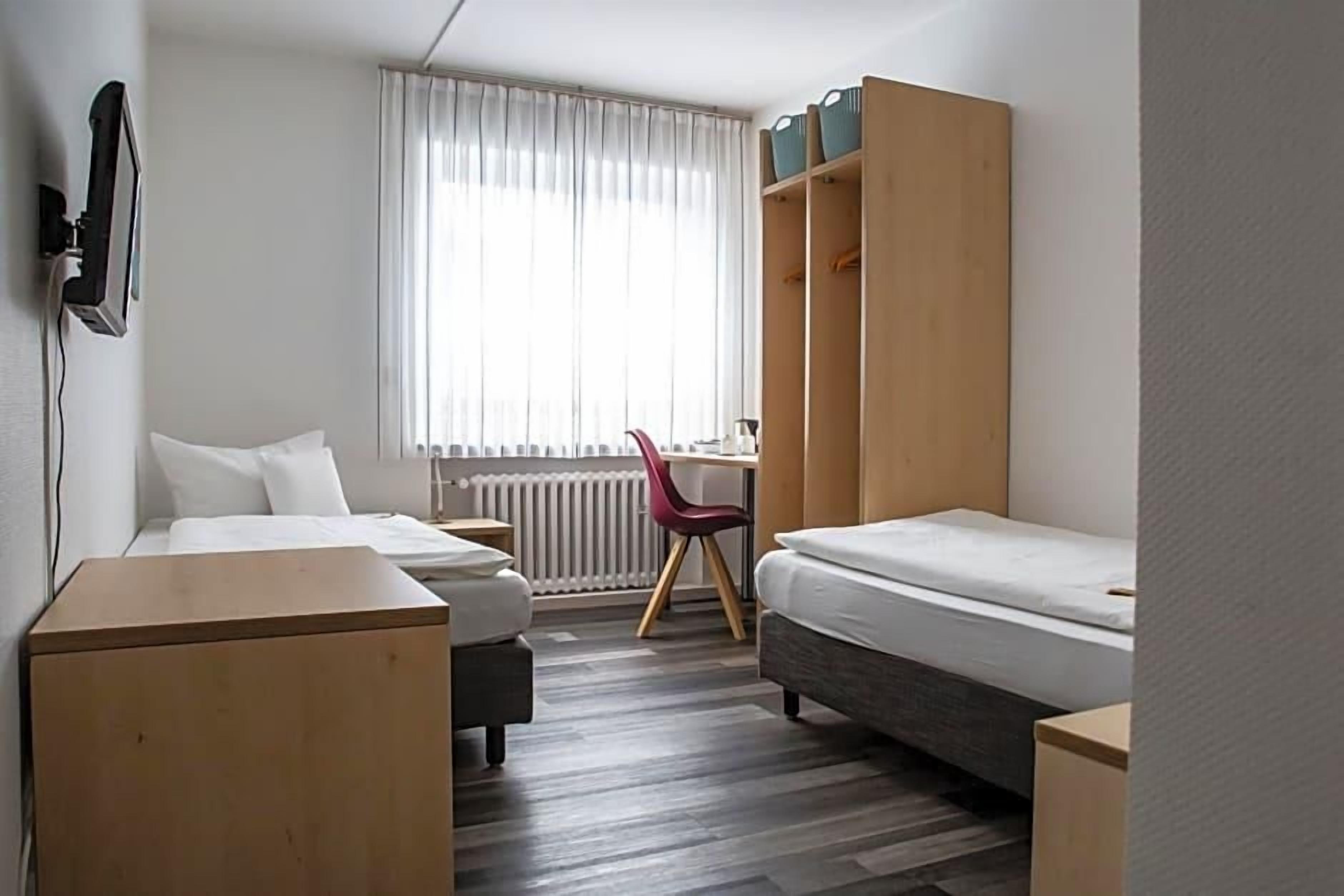 A compact hotel room with two beds, a desk, a chair, and a wardrobe.