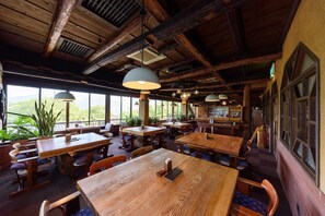 Un interior rústic de fusta amb diverses taules i bancs de fusta, grans finestres que ofereixen vistes a la vegetació, i llums penjants.