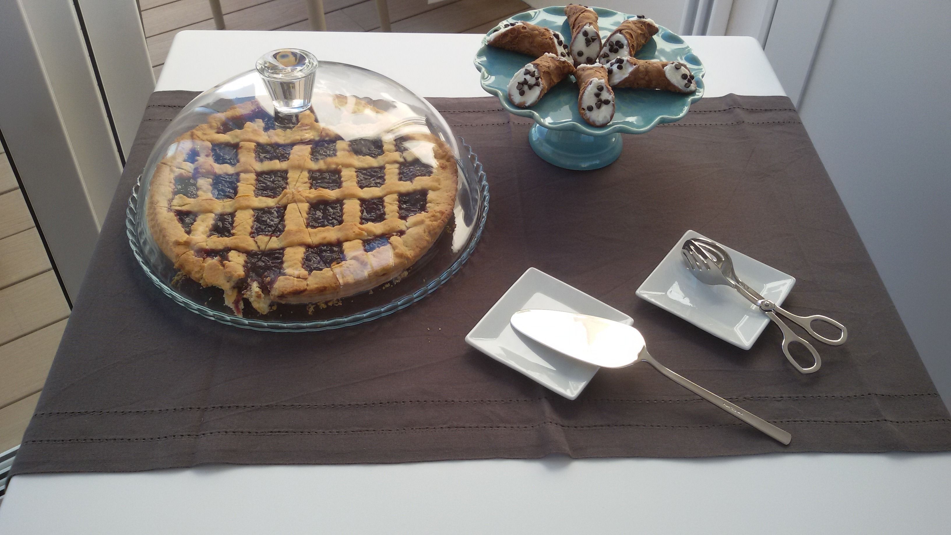 A pie on a glass plate with a lattice crust, a plate with a fork and scissors, and a blue plate with cannoli.