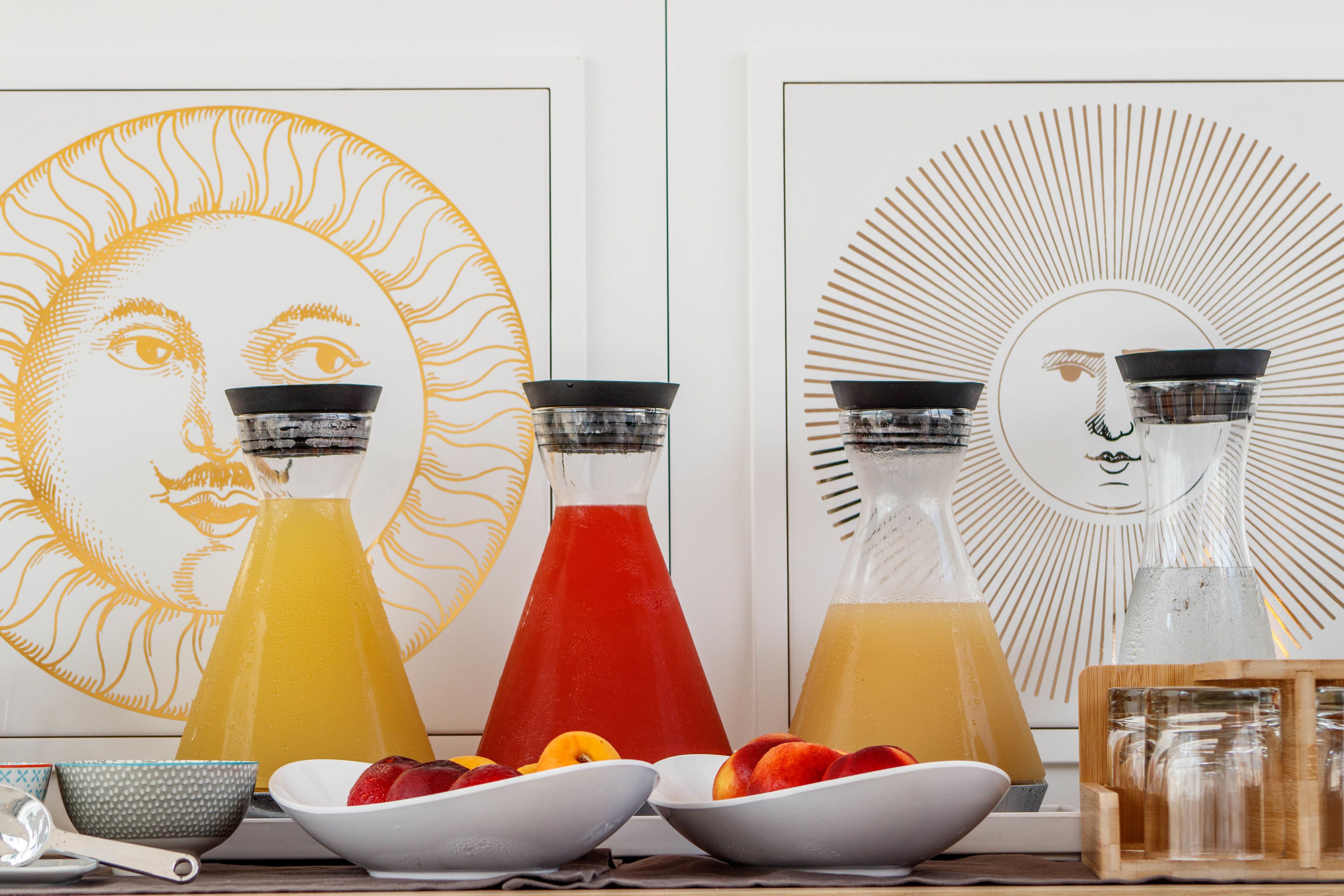 A table with three glass bottles of different colored beverages, a bowl of fruit, and a decorative wall with a sun design.