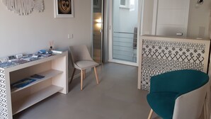 A modern living room with a desk, a chair, and a teal armchair. There is a door leading to a balcony with a railing.