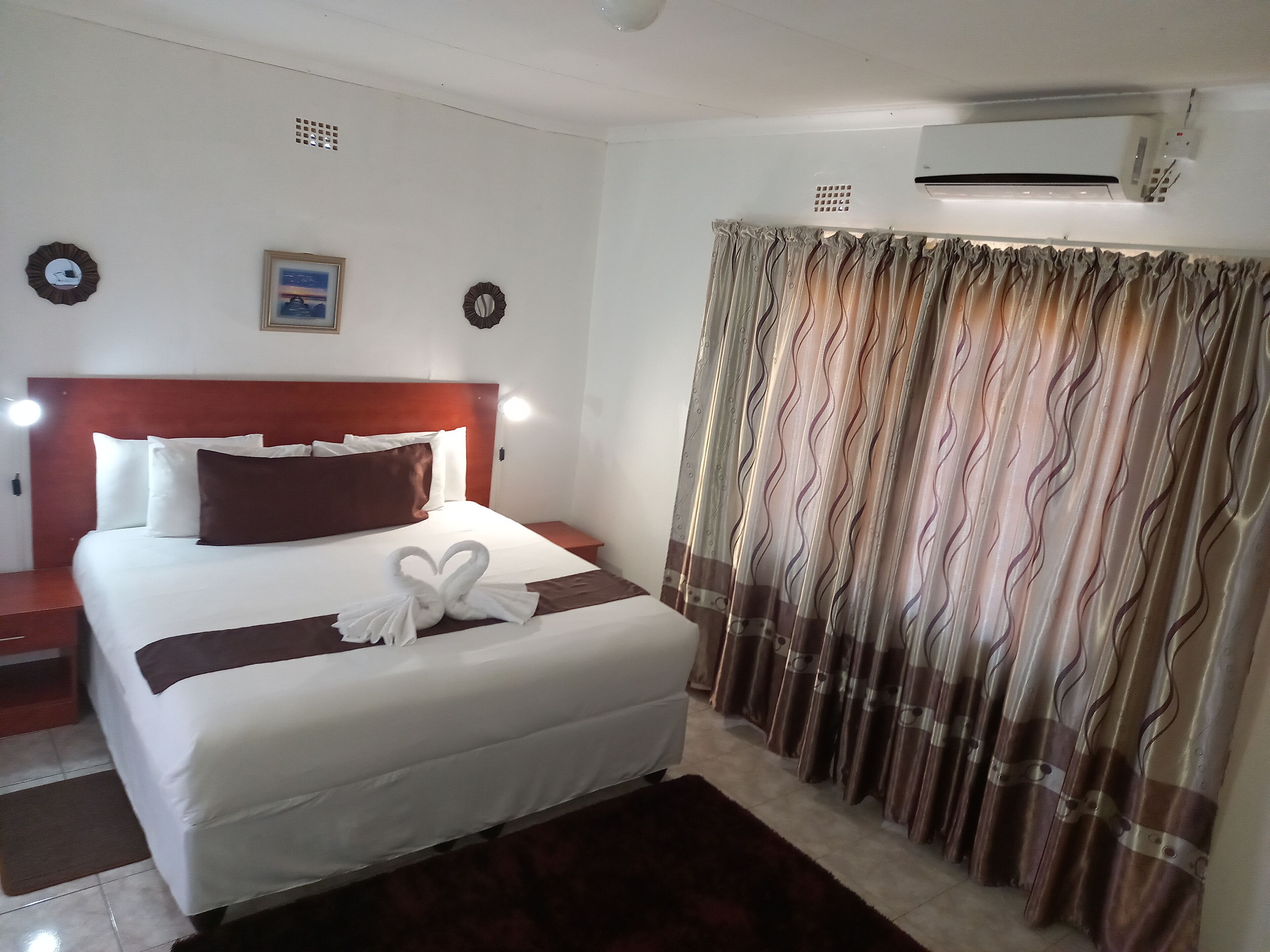 A hotel room with a neatly made bed, a headboard, a bedside table, a wall-mounted air conditioner, and a curtain with a wavy pattern.