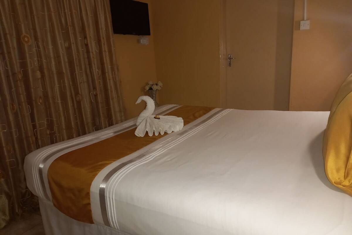 A hotel room with a neatly made bed, a swan-shaped towel, a vase with flowers, and a television mounted on the wall.