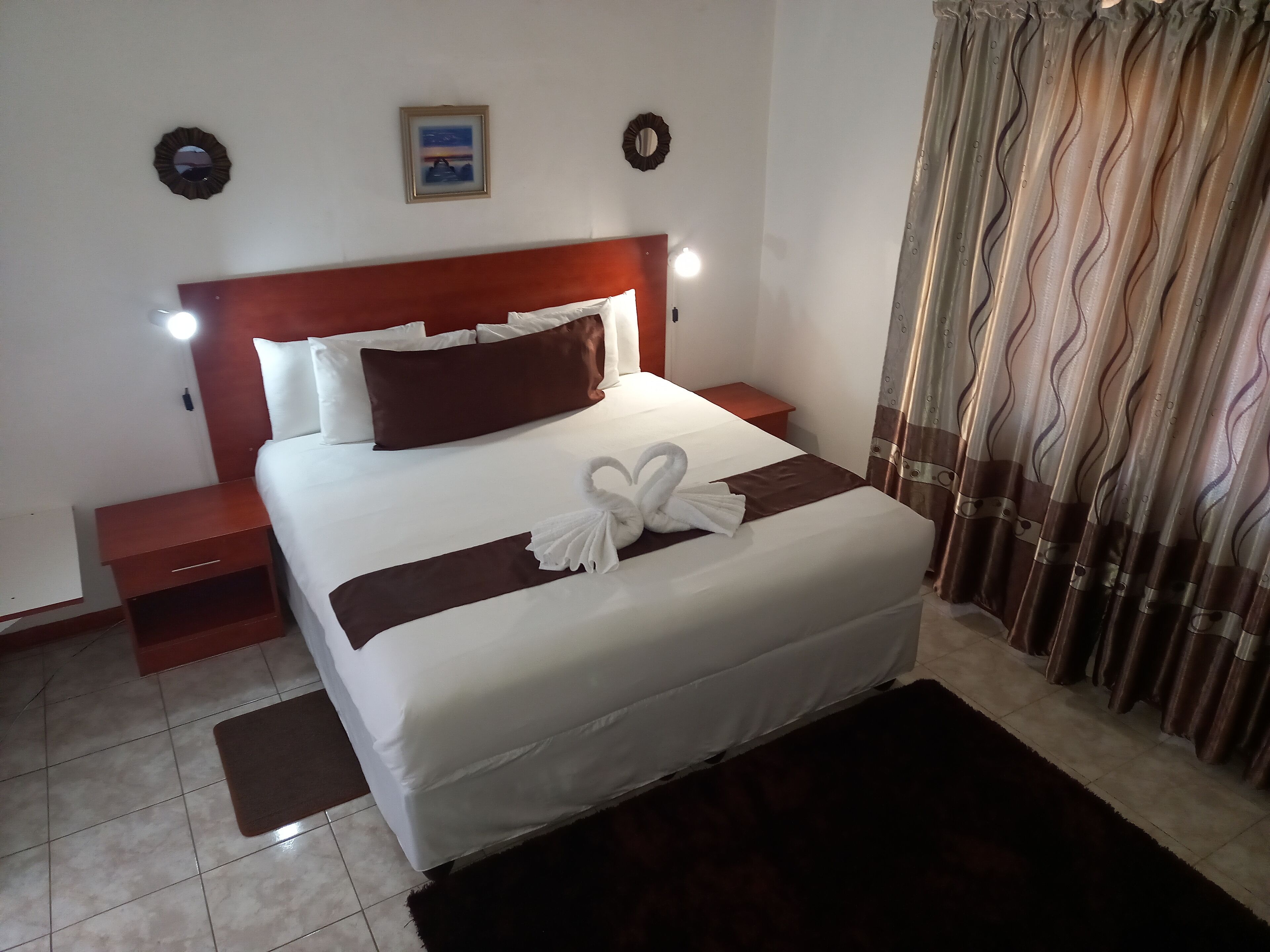 A neatly made bed with white and brown bedding, a headboard with integrated reading lights, a small bedside table, and a decorative towel on the bed.