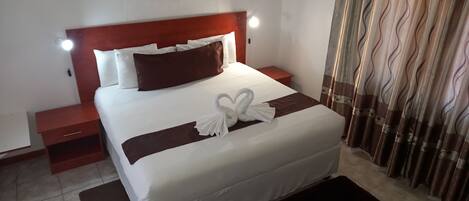 A neatly made bed with white and brown bedding, a headboard with integrated reading lights, a small bedside table, and a decorative towel on the bed.
