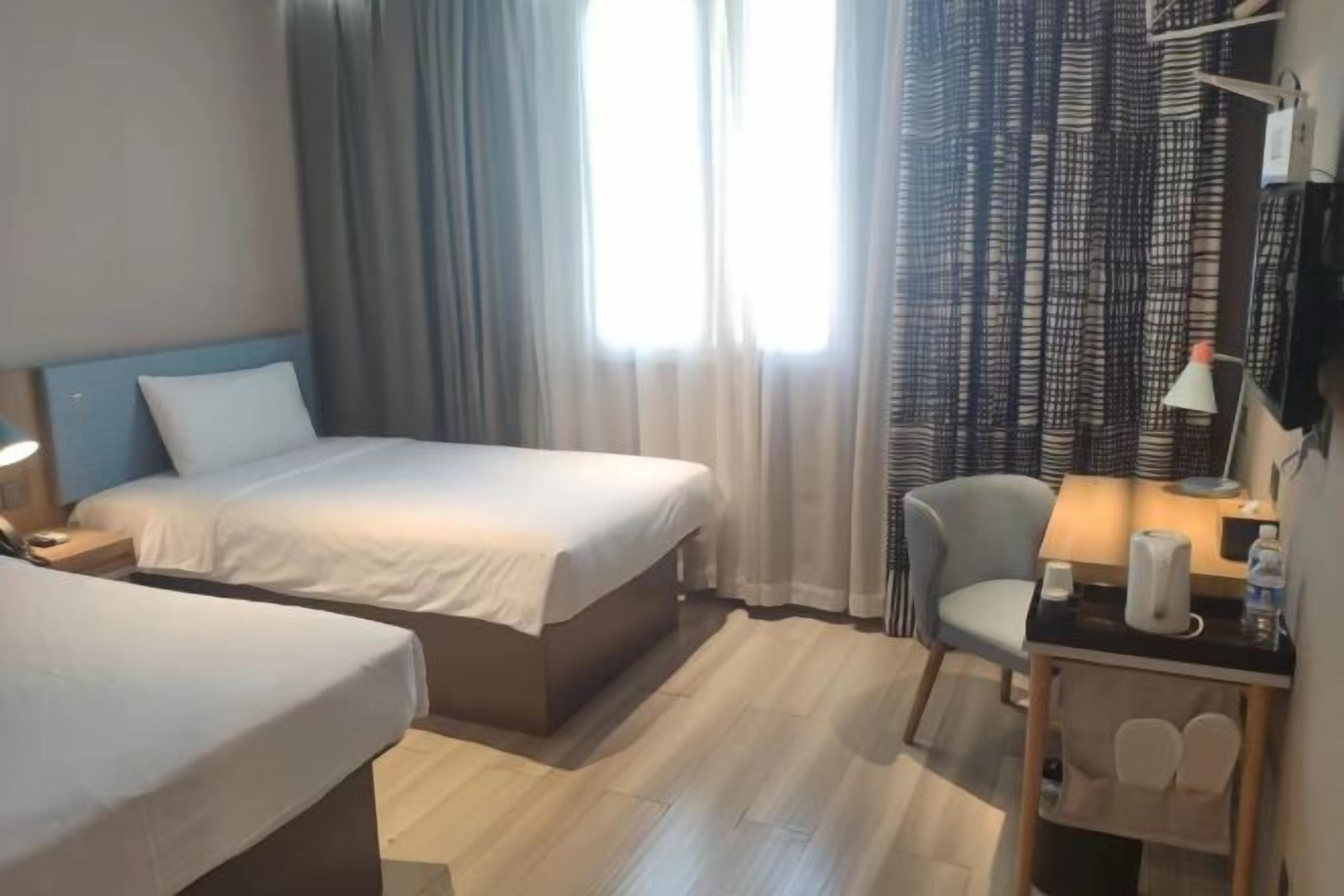 Una habitación de hotel con cama, una silla, un escritorio con lámpara y una ventana con cortinas.