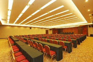 Un salón de conferencias con filas de mesas y sillas, un escenario y suelo alfombrado.