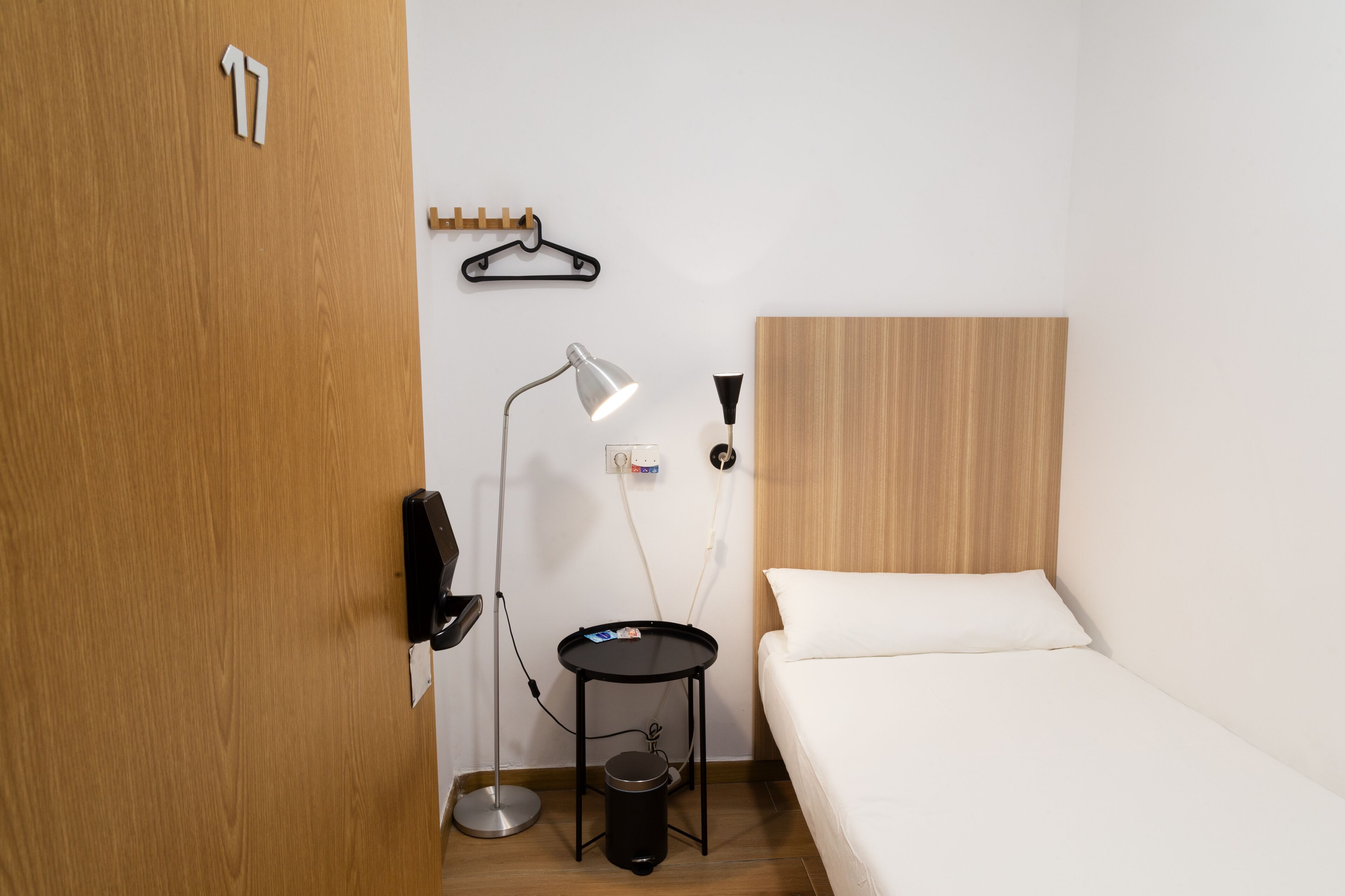 A small, neatly organized room with a wooden door, a bed with white bedding, a small round table with a lamp, and a coat hook on the wall.