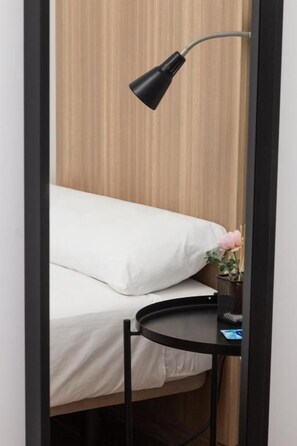 A bed with white bedding, a small round table with a black lamp, and a potted plant with pink flowers. - Siesta & Go - Hostel (Madrid)