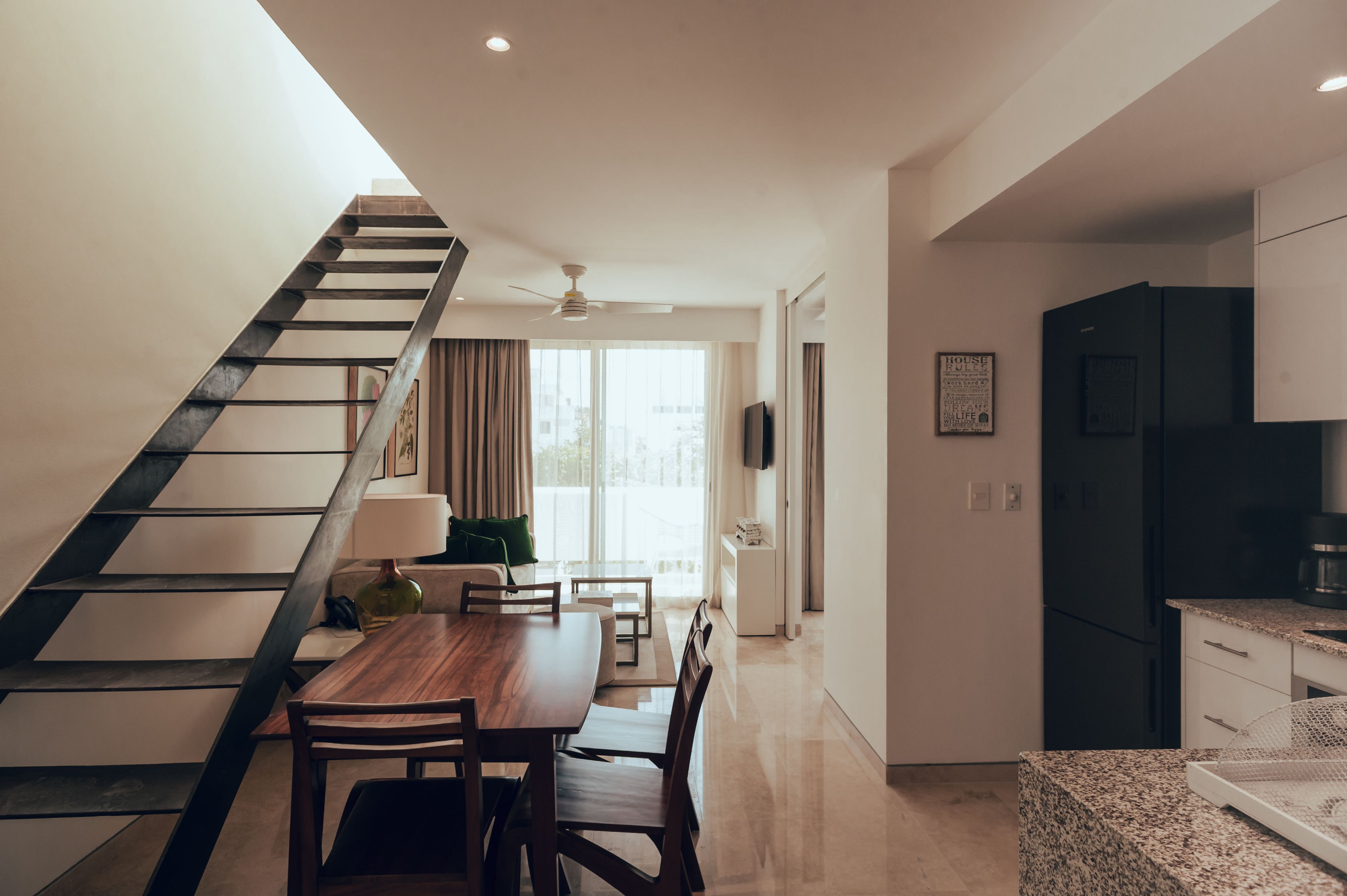 A modern kitchen with a black refrigerator, a dining area with a wooden table and chairs, and a staircase with a metal railing.