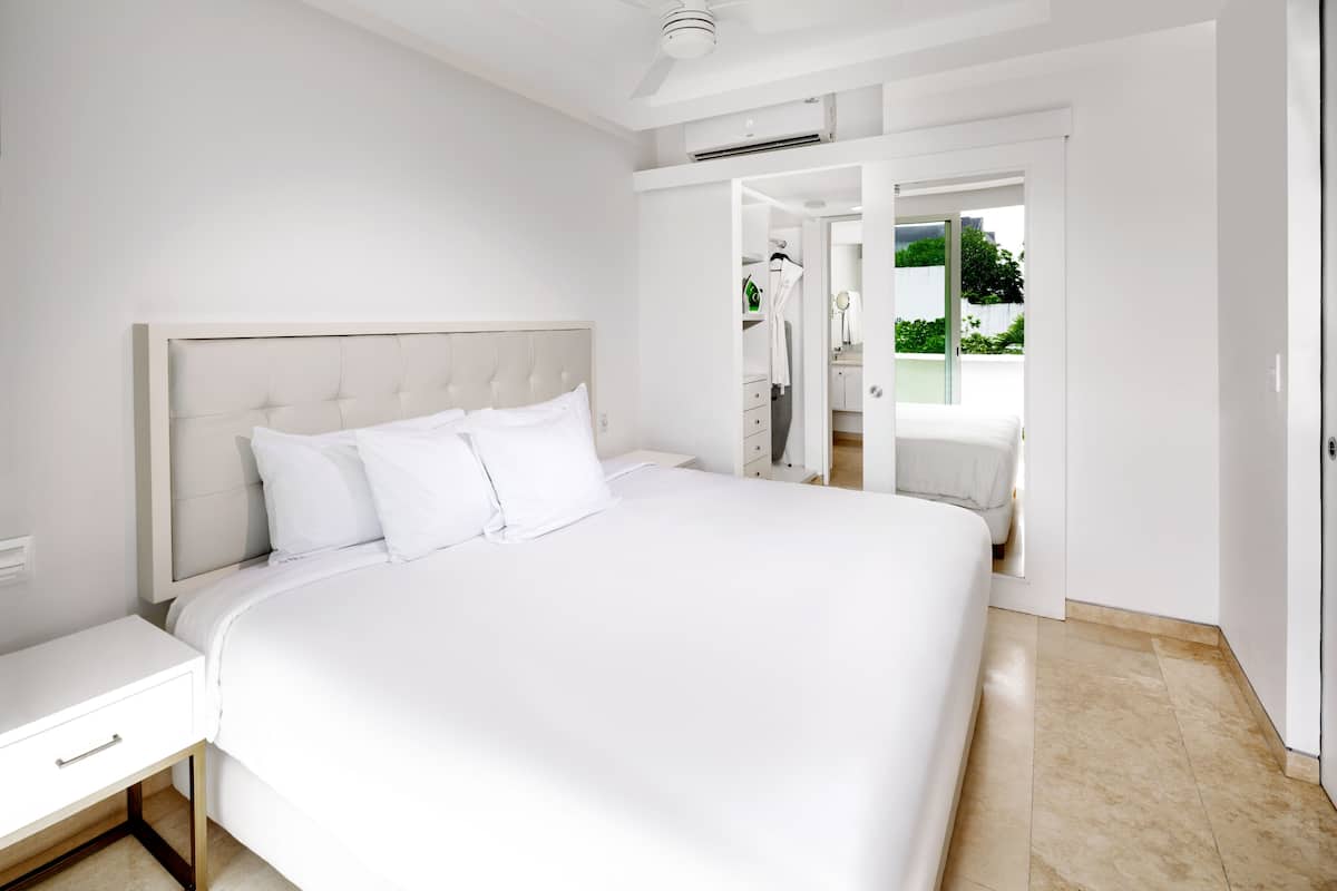 A large bed with a tufted headboard in a bright, white room with a view of a garden through an open door.