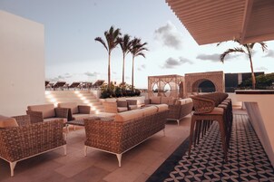 A rooftop lounge area with wicker furniture, palm trees, and a patterned floor.