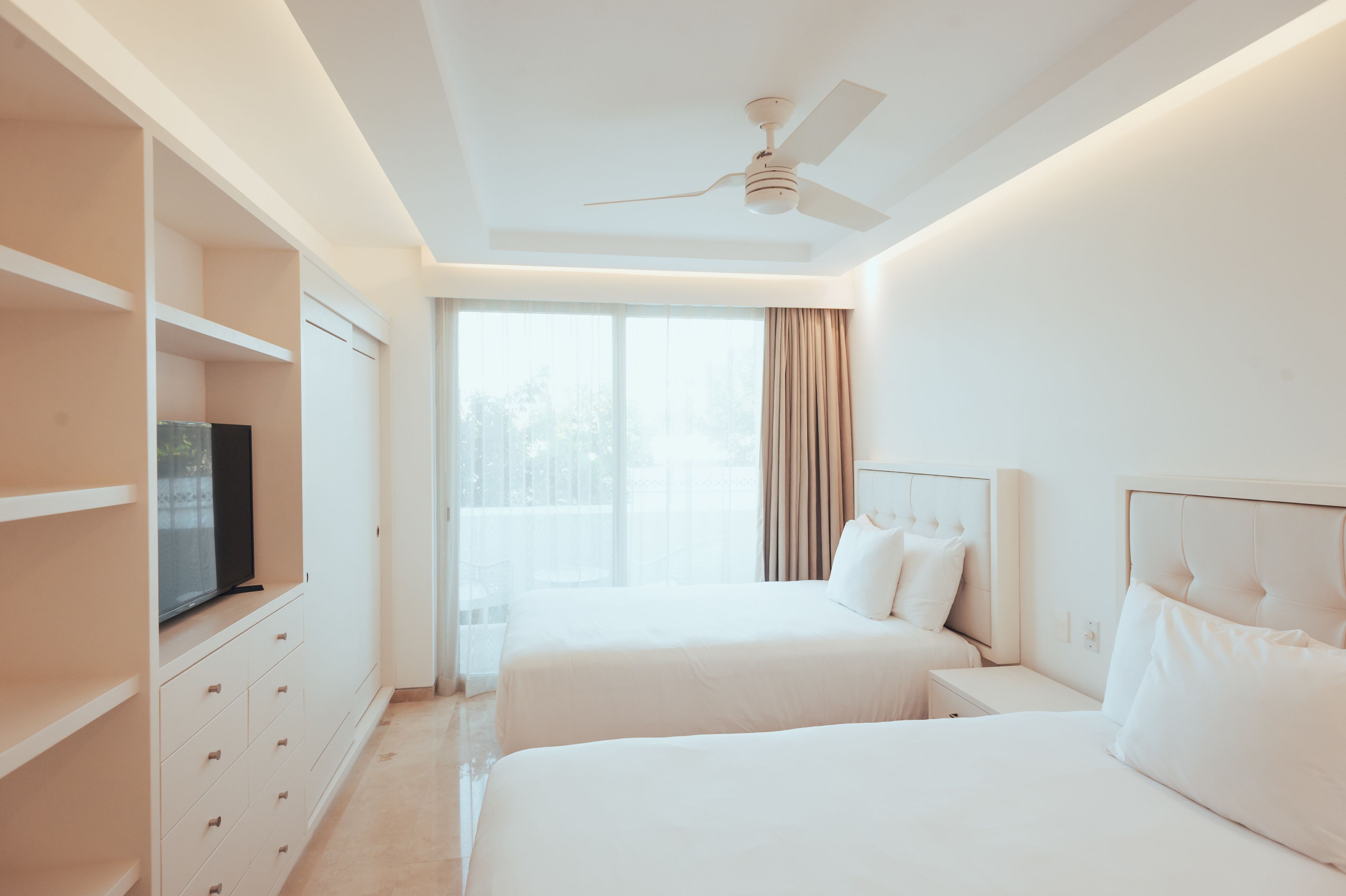 A hotel room with two beds, a TV on a cabinet, a ceiling fan, and a large window with curtains.