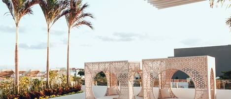 A rooftop lounge with wicker furniture, a bar, and palm trees in the background.